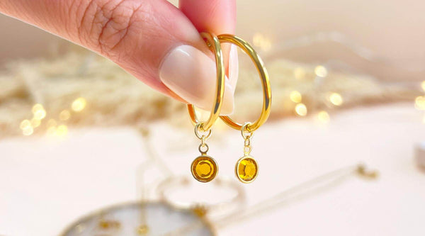 Image shows close up of gold hoops with the yellow November topaz birthstone in front of a soft cream backdrop.