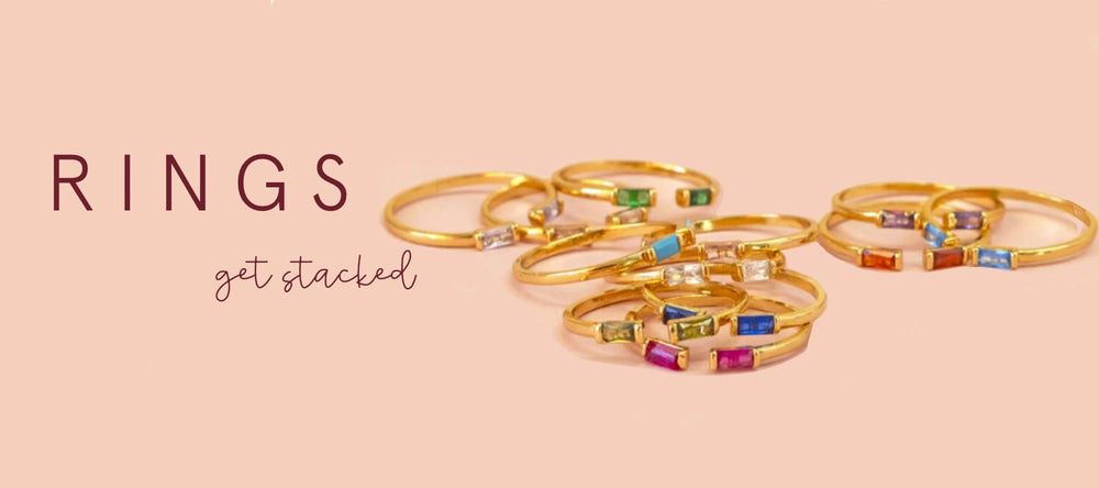 Image shows gold birthstone rings on a pink background the the wording rings get stacked