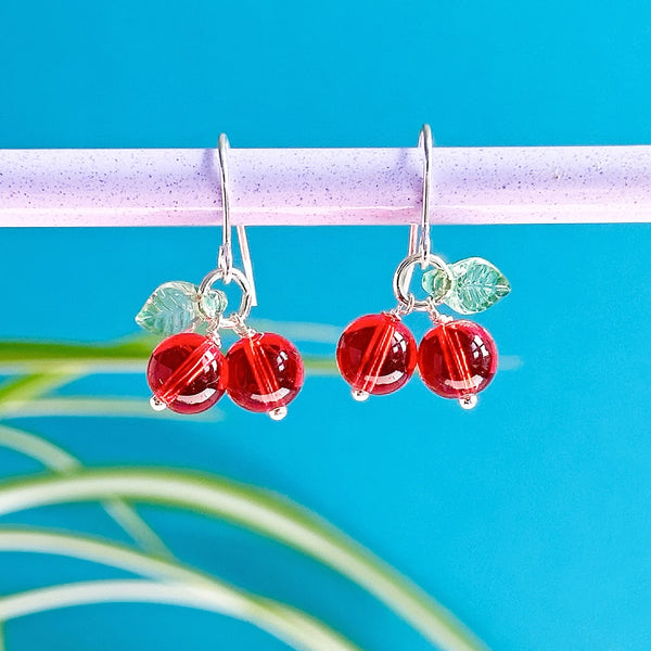 Im age shows a pair of silver plated glass cherry earrings hanging with a blue backdrop