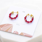 Image shows silver July Birthday Ruby Birthstone Beaded Huggie Earrings lying on a July birthstone characteristic card