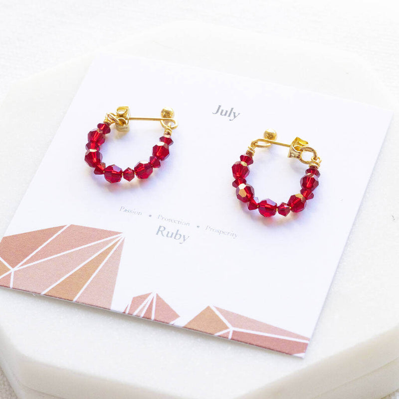 Image shows silver July Birthday Ruby Birthstone Beaded Huggie Earrings lying on a July birthstone characteristic card