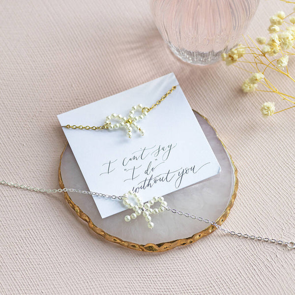 Image shows gold plated Bridesmaid Pearl Bow Bracelet on an "I can't say 'I do' without you" sentiment card. Silver plated Bridesmaid Pearl Bow Bracelet sits on a crystal jewellery display.