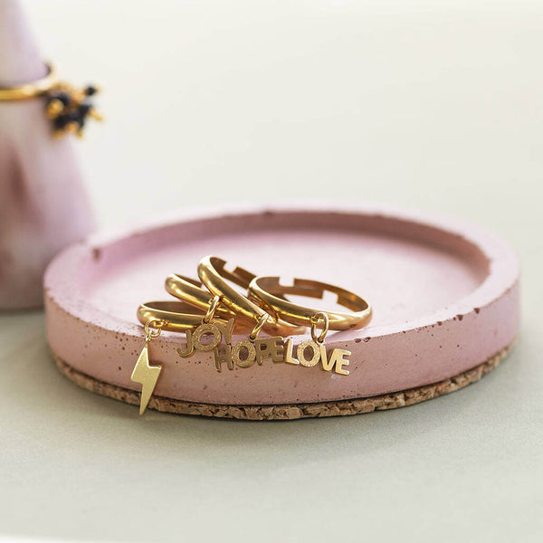 Love, Joy, hope and lighting bolt gold plated adjustable charm rings stacked on a pink jewellery trinket plate.
