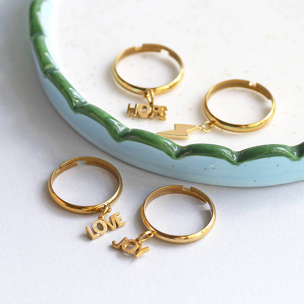 Love, Joy, hope and lighting bolt gold plated adjustable charm rings set on a jewellery trinket plate.
