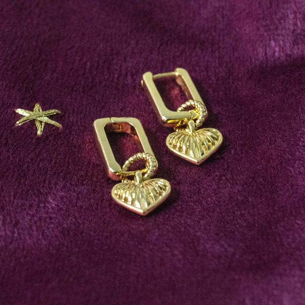 Image shows a pair of Ribbed Heart Huggie Earrings on a  maroon backdrop