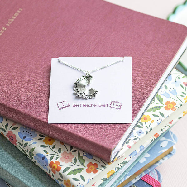 A silver plated necklace with a pencil shaving charm displayed on a Best Teacher Ever sentiment card sat on top of a selection of journals and notebooks.