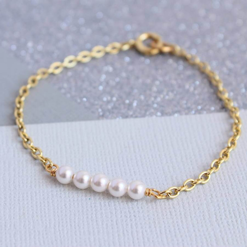 Image shows delicate Swarovski pearl bracelet in gold plated brass, with five dainty white pearls. on a grey and cream backdrop.
