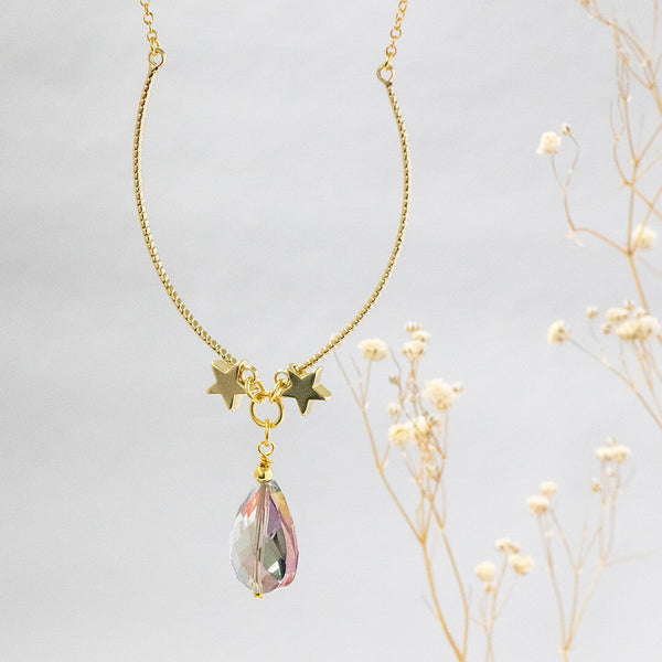 Image shows gold plated Elysium necklace suspended in front of a grey background. A gorgeous blue iridescent crystal suspended between two gold stars.