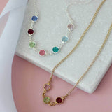 Image shows two necklaces on a pink and white background. Silver necklace seven birthstones; September Sapphire, July Ruby, August Peridot, October Rose, May Emerald, March Aquamarine, June Light Amethyst. Gold necklace has three birthstones, April Crystal, August Peridot and January Garnet. More birthstones can be added.