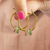 Model holds pair of gold plated birthstone hoop earrings on a white backdrop. Birthstone crystal rings from left to right: October Rose, March Aquamarine, May Emerald.