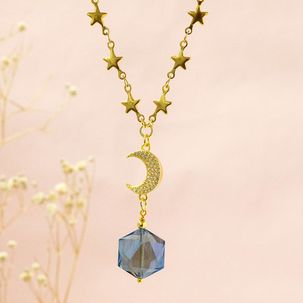 Image shows Galaxy Necklace with a gorgeous blue crystal, crystal encrusted crescent moon, and gold dainty star chain detail. Necklace is suspended in front of a pink backdrop. 