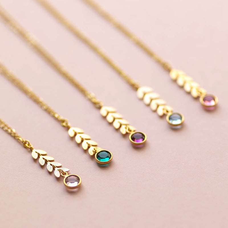 Image shows five gold leaf vine necklaces on a pink backdrop. Each with a different Swarovski birthstone. From left to right: April Crystal, May Emerald, February Amethyst, March Aquamarine, October Rose.