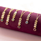Image shows six gold leaf vine necklaces drape on a burgundy stand andon a pink backdrop. Each with a different Swarovski birthstone. From left to right: September Sapphire, April Crystal, May Emerald, February Amethyst, March Aquamarine, October Rose.