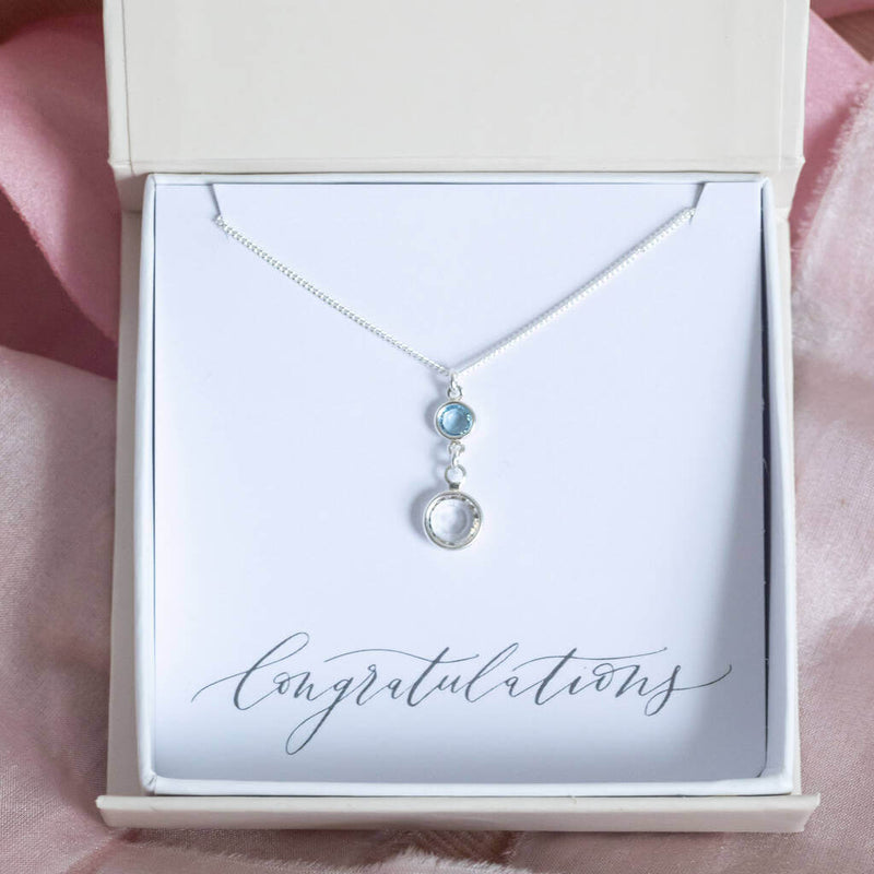 A sterling silver chain pendant with a smaller birthstone in March aquamarine and the larger birthstone to represent mother is April crystal. Displayed on a 'congratulations' sentiment card in a JOY gift box.