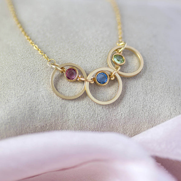 Image shows Triple Linked Circle Swarovski Birthstone Necklace on a nude jewellery pillow. Birthstones from left to right in selection order: October Rose, September Sapphire and August Peridot.