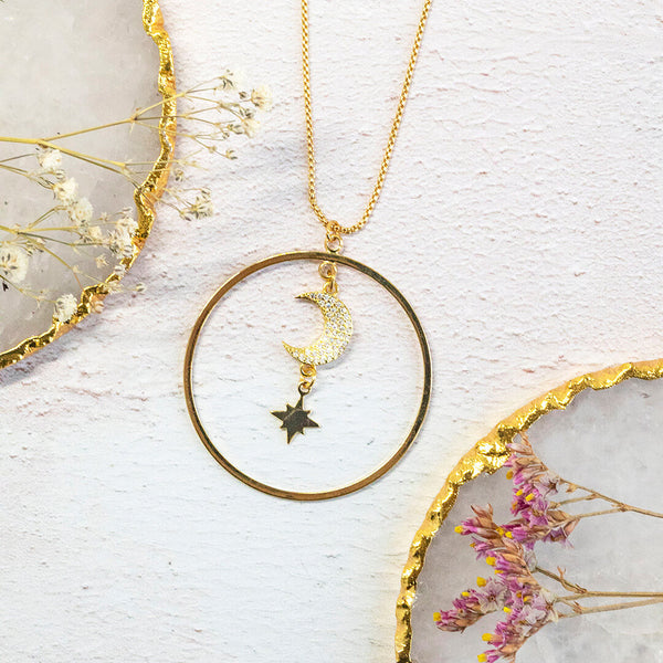 Image shows Luna Gold plated necklace on a white flat lay. Necklace comprises of a golden circle, suspended within this is a sparkling crescent moon and star charm. 