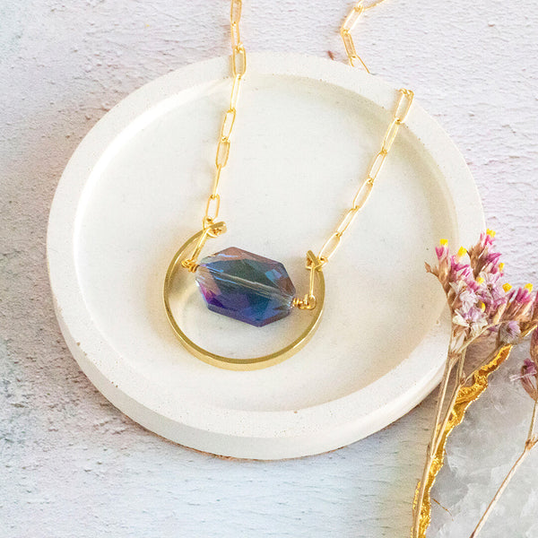 Image shows supernova blue crystal pendant necklace with blue crystal and golden arc on a white and floral backdrop.