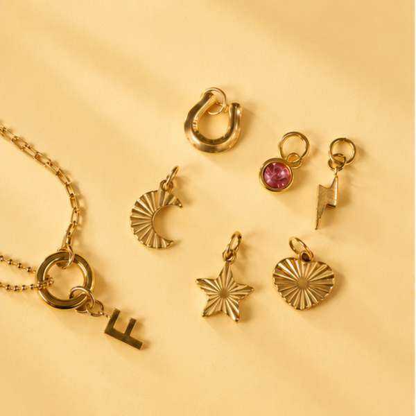 Personalised Week Of Charms Necklace in gold with interchangeable initial, birthstone, moon, star, heart, lightning bolt and horseshoe charms on a soft cream background.