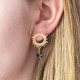 Image shows a model with blonde hair wearing a pair of gold sun earrings with black star detail