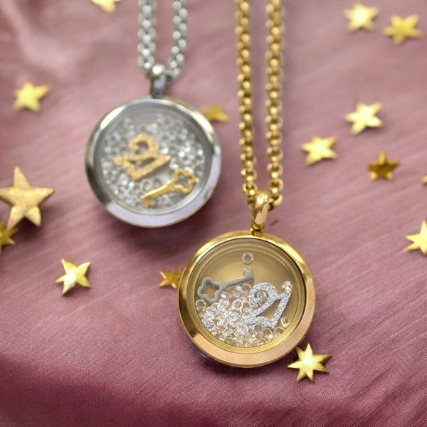 Image shows two 21st birthday locket necklaces, one in silver and one in gold on a dusky pink fabric background with scattered gold stars