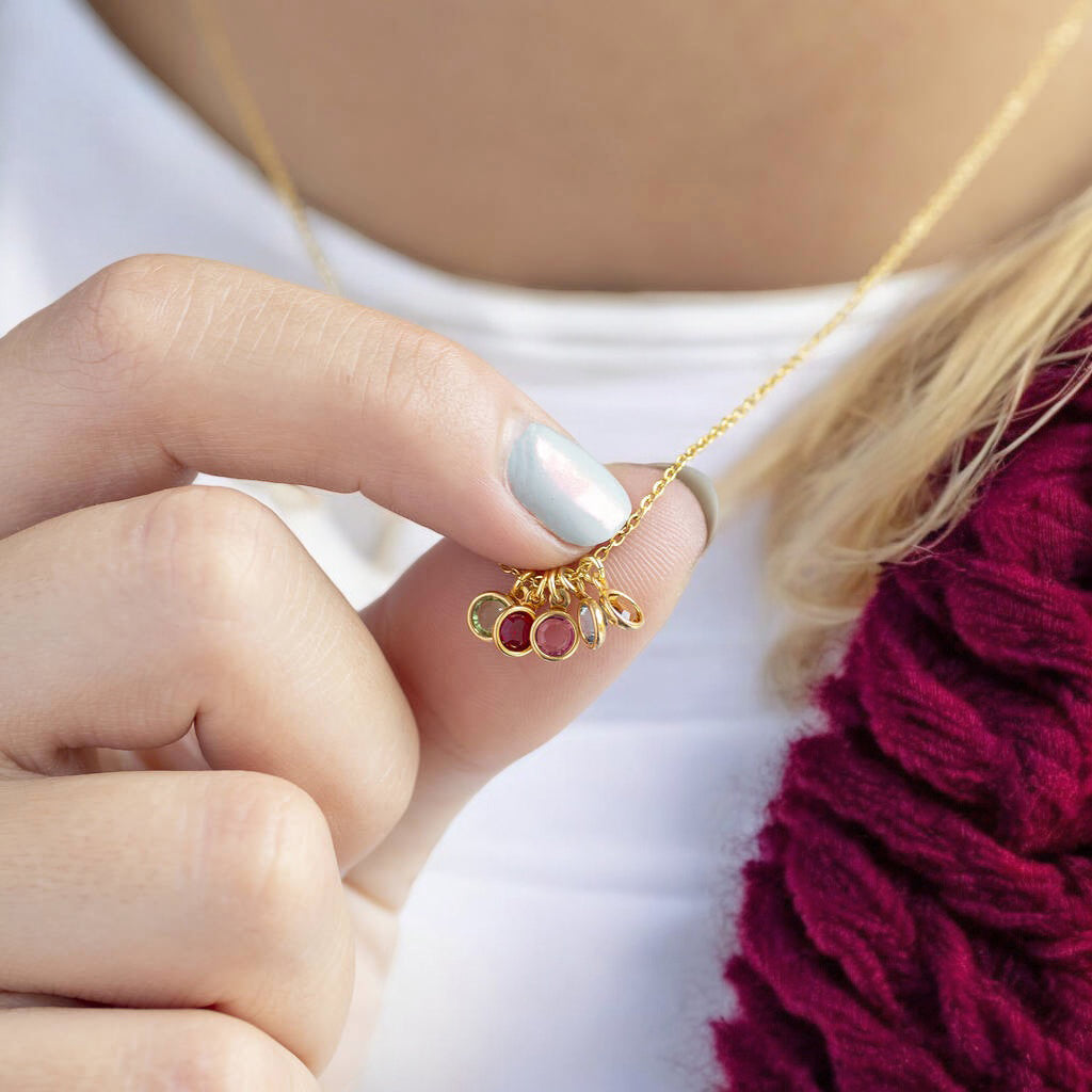 Image shows a woman in a burgundy cardigan holding onto a gold necklace  with mini fa,ily birthstones that is around her neckwith 