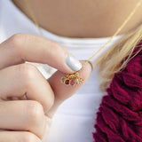 Image shows a woman in a burgundy cardigan holding onto a gold necklace  with mini fa,ily birthstones that is around her neckwith 