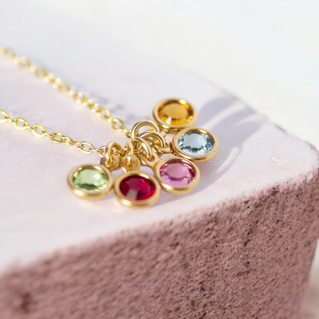 Image shows a gold mini family birthstone necklace on a pink concrete slap