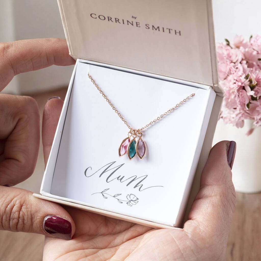 Image shows a woman's hand holding a gift box that reads mum upon which there is a rose gold family birthstone necklace