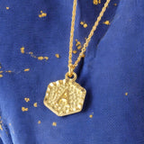 Image shows a gold hexagon initial A necklace on a deep blue and gold background