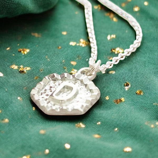 image shows a silver hexagon initial necklace with the letter D on a green and gold background