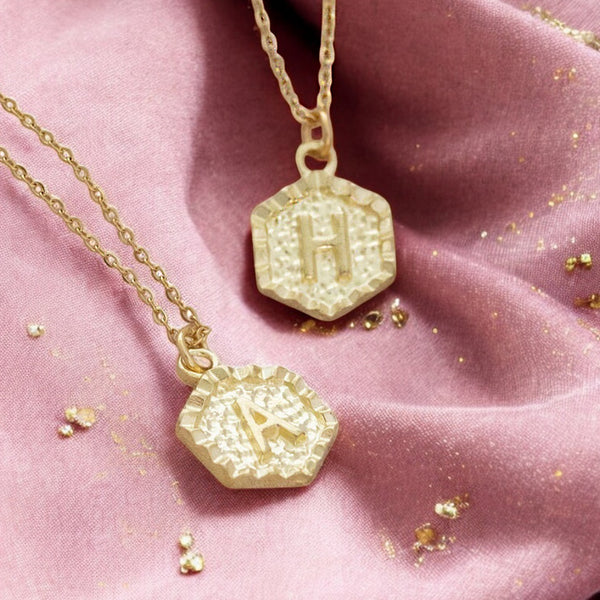 image shows two gold hexagon initial necklaces on a dusky pink fabric background