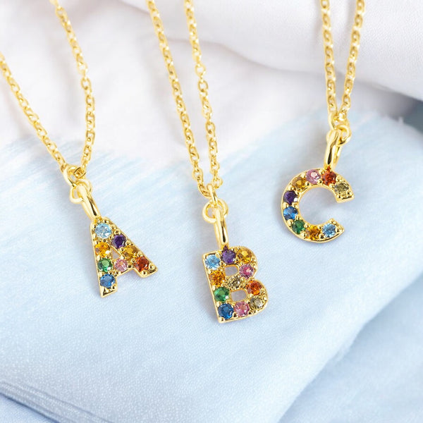 Image shows three gold necklaces with letters A B and C that are set with multi coloured gemstones son a pale blue fabric background