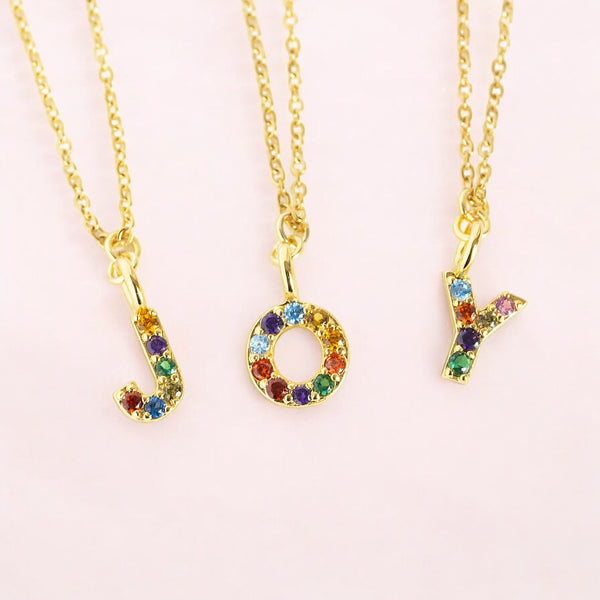 Image shows three gold necklaces with letters that are set with multi coloured gemstones spelling out the word joy on a pale pink background