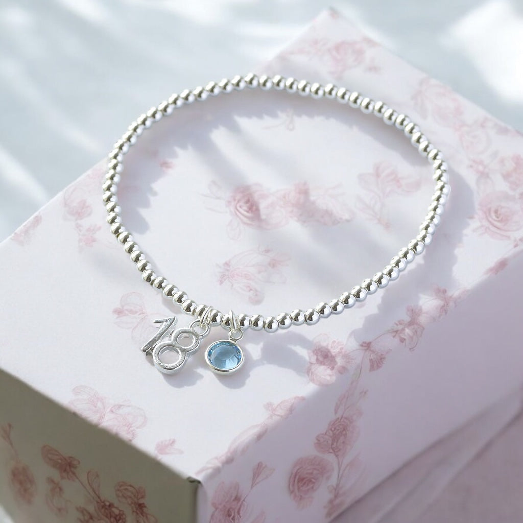 Image shows a silver beaded 18th birthday charm bracelet wiuth march birthstone on a pink floral jewellery box