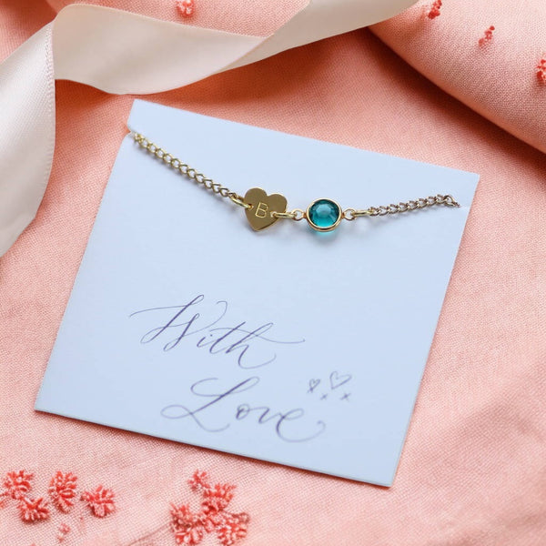 image shows a personalised heart and birthstone bracelet on a white card that reads with love on a coral background