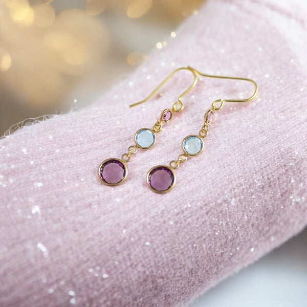 image shows a pair of gold three generations birthstone earrings on a sparkly mauve fabric scarf with twinkly lights in the background