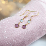 image shows a pair of gold three generations birthstone earrings on a sparkly mauve fabric scarf with twinkly lights in the background
