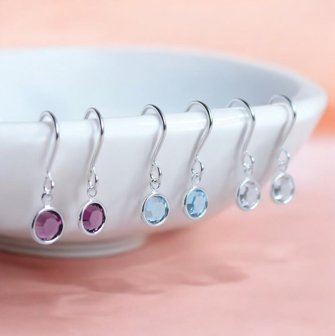 Images show february, march and april crystal birthstone earrings hanging from the edge of a white trinket dish