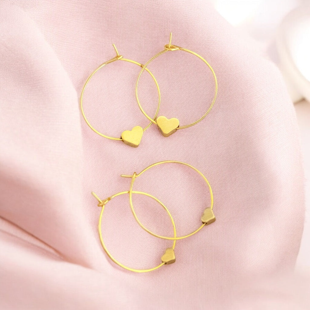 Image shows two pairs of gold hoop earrings with heart detail on a pale pink fabric background