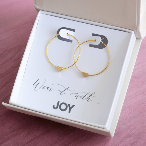 Images shows a gift box containing a pair of gold heart hoop earrings on a card that reads wear it with joy