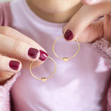 Image shows a woman in a pink top holding a pair of gold hoop earrings with tiny heart detail