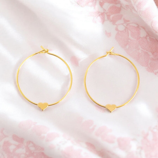 Image shows a pair of gold heart hoop earrings on a pale pink and white background