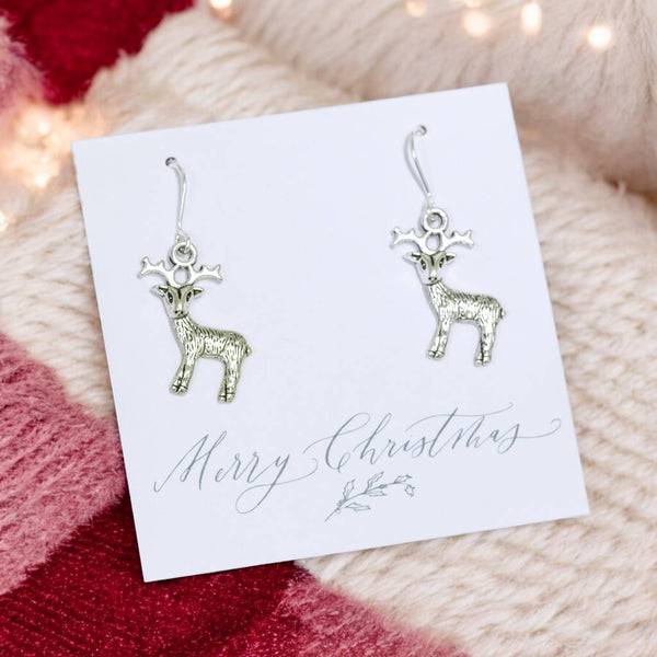 Image shows a pair of reindeer earrings on a white merry christmas card on a pink and cream wool background