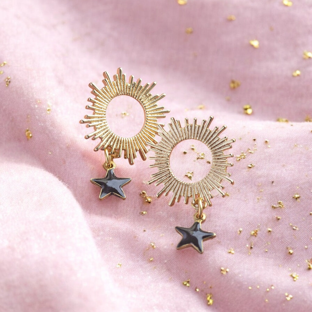 images shows a pair of gold sun earrings with black enamel star charm detail on a pale pink sparkly background