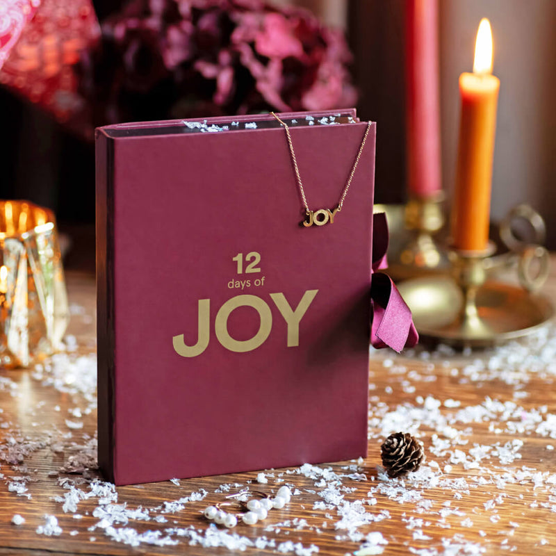Image shows the 12 Days of JOY Advent Calendar with a golden JOY necklace draped over the front. Background features a lit candle and faux snow.