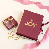 Image shows the 12 Days of JOY burgundy jewellery advent calendar, gold miscellaneous jewellery and a small wrapped gift with a golden bow (not included) sitting on a pink backdrop with a dark pink ribbon.