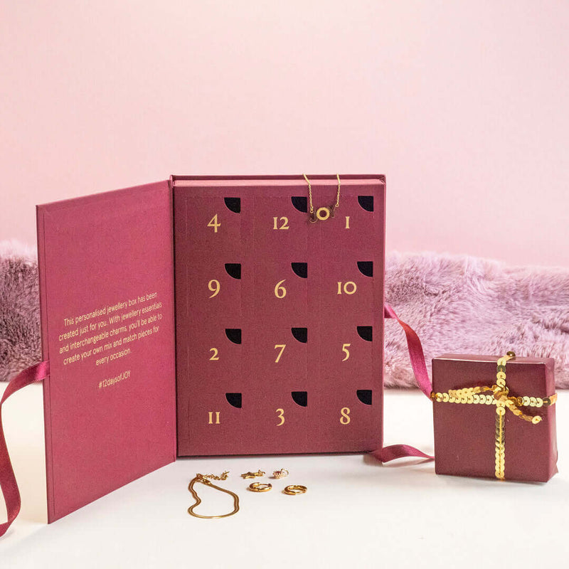 Image shows a JOY burgundy jewellery advent calendar that is open to display gold numbers, displayed on a pink backdrop with a gold necklace and gold earrings on the table in front of the calendar, and a small wrapped gift with a golden bow (not included).  Edit alt text