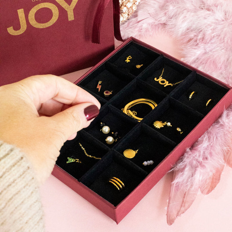 Image shows the jewellery insert for the 12 Days of JOY Advent Calendar filled with gold jewellery. Models hand is reaching for the necklace. Pink backdrop features pink feathers.