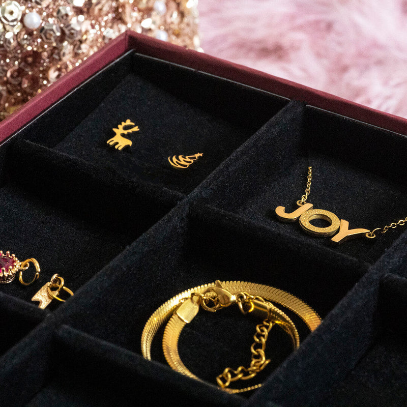 Image features close up of black jewellery insert with a pair of Christmas earrings, JOY necklace and flat snake chain bracelet. Blurred background features gold sequins and pink feathers.