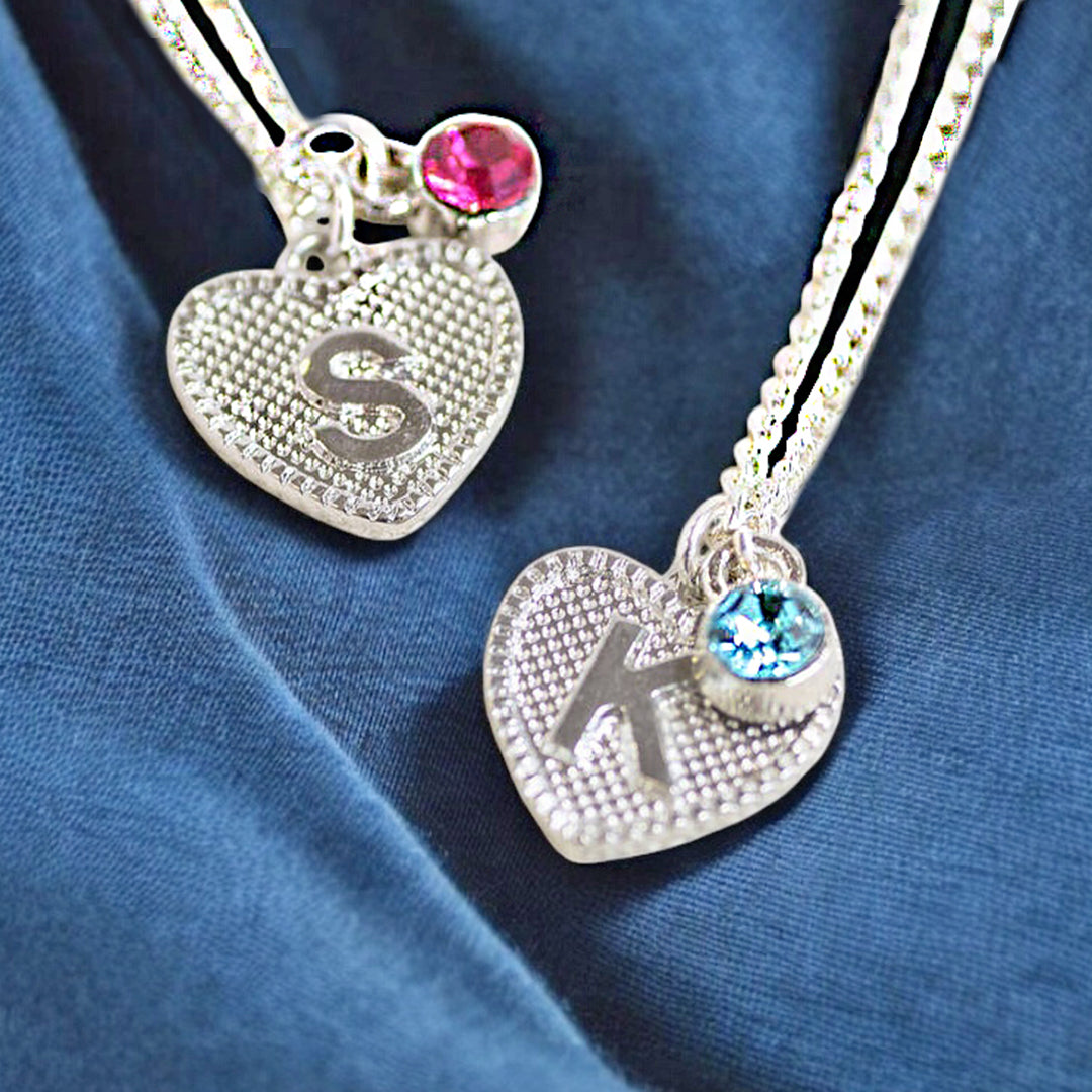 image shows two children silver initial heart necklaces with birthstone detail on a deep blue fabric background